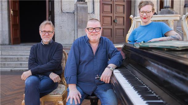 Drei Personen, zwei Männer und eine Frau, an einem Klavier.