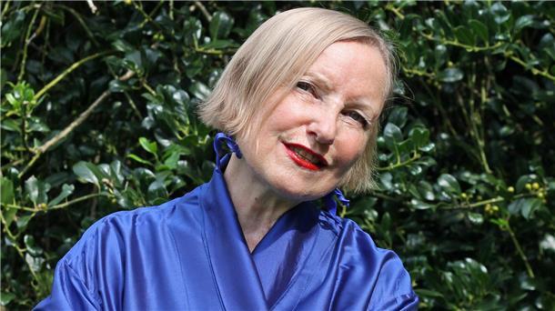 Eine Frau in einem blauen Outfit vor grünem Laub, lächelnd mit rotem Lippenstift. Es ist Anna Haentjens, die im TiF Bremerhaven auftreten wird - mit einer Hommage an Lale Andersen. 