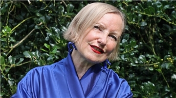 Eine Frau in einem blauen Outfit vor grünem Laub, lächelnd mit rotem Lippenstift. Es ist Anna Haentjens, die im TiF Bremerhaven auftreten wird - mit einer Hommage an Lale Andersen. 
