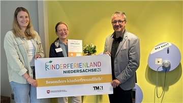 Drei Personen halten eine Urkunde und ein Banner mit der Aufschrift „Kinderferienland Niedersachsen – Besonders kinderfreundlich!“ vor einer gelben Wand.