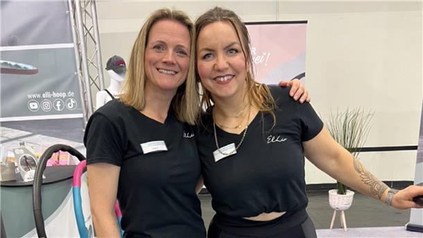 Zwei Frauen stehen lächelnd nebeneinander an einem Messestand, tragen schwarze T-Shirts mit Namensschildern und dem Aufdruck „Elli“. Im Hintergrund sind Hula-Hoop-Reifen und Werbematerialien zu sehen.