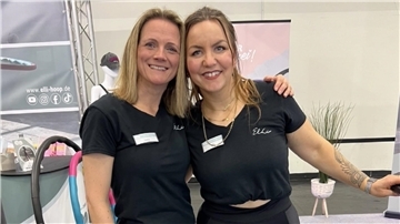Zwei Frauen stehen lächelnd nebeneinander an einem Messestand, tragen schwarze T-Shirts mit Namensschildern und dem Aufdruck „Elli“. Im Hintergrund sind Hula-Hoop-Reifen und Werbematerialien zu sehen.
