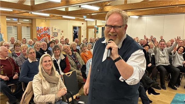 Lars Cohrs bringt 180 Loxstedter mit seiner Weihnachtslesung in Stimmung Ein Mann mit Brille, Bart und Mikrofon steht lachend im Vordergrund eines Saals voller Menschen.