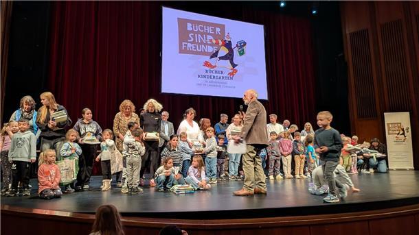 Kinder und Erwachsene stehen auf einer Theaterbühne. Im Hintergrund ist eine Leinwand mit dem Logo „Bücher sind Freunde“ zu sehen.