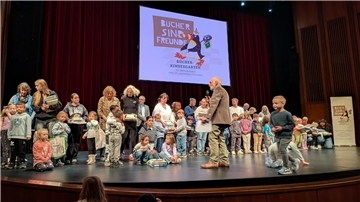 Kinder und Erwachsene stehen auf einer Theaterbühne. Im Hintergrund ist eine Leinwand mit dem Logo „Bücher sind Freunde“ zu sehen.