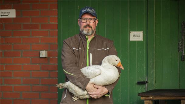 Ein Bauer steht vor einem grünen Tor und hält eine Gans im Arm.
