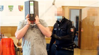 Der Angeklagte verbirgt sein Gesicht hinter einem Aktenordner, als er zum Prozessauftakt von einem Justizvollzugsbeamten an seinen Platz geführt wird. Schoener