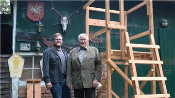 Zwei Männer stehen vor einem grünen Gebäude mit Jagddekorationen. Neben ihnen steht ein neu gebauter hölzerner Hochsitz.