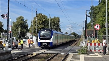 Personenzug fährt über einen Bahnübergang, rechts und links stehen Arbeiter