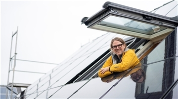 Ein Mann mit Brille und gelber Jacke lehnt sich lächelnd aus einem geöffneten Dachfenster. Das Dach ist mit glänzenden Solarpaneelen bedeckt. Im Hintergrund ist ein Baugerüst zu sehen.