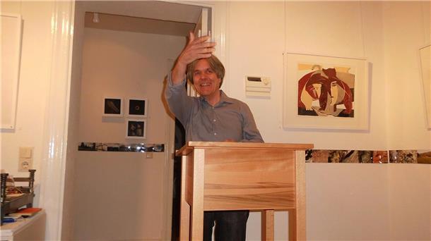 Ein Mann steht an einem Rednerpult und hebt grüßend die Hand. 