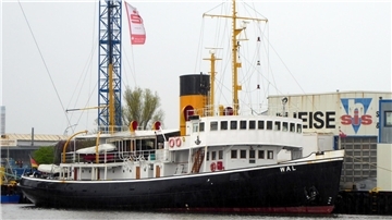 Der historische Dampfeisbrecher „Wal“ liegt im Hafen vor einer Werftanlage. Das Schiff ist schwarz-weiß mit einem markanten gelb-schwarzen Schornstein. Rettungsringe hängen an der Reling, und im Hintergrund sind Kräne sowie industrielle Gebäude mit Werbeaufschriften zu sehen.