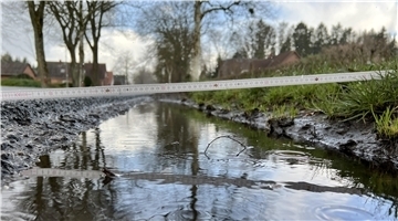 Aufgefahrener Seitenraum einer Landesstraße, in dem sich Wasser sammelt Von links nach rechts liegt ein Zollstock. Der Seitenraum liegt höher als die Straße.