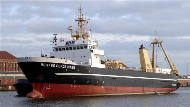 Der Hochseetrawler „Gerda Maria“ mit der Kennung ROS 786 fährt auf dem Wasser, im Hintergrund sind Hafengebäude und ein wolkenverhangener Himmel zu sehen.