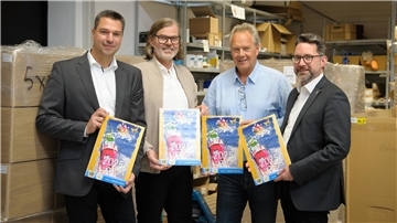 Vier Männer mit jeweils einem Adventskalender in der Hand stehen in einem Lager vor Kartons.