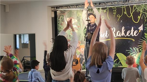 Ein Mann steht auf einer Bühne und hebt die Hände, mehrere Kinder stehen davor.