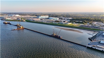 Luftaufnahme vom Hafen in Cuxhaven mit mehreren Bauschiffen und Großgeräten, die an einer langen Spundwand im Wasser arbeiten. Im Hintergrund sind Lagerhallen, Industrieanlagen und geparkte Fahrzeuge zu sehen.