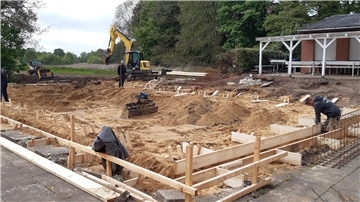 Baustelle mit Bagger, Bauarbeitern und Holzverschalung vor einem Vereinsheim; der Boden ist ausgehoben und für ein neues Fundament vorbereitet.