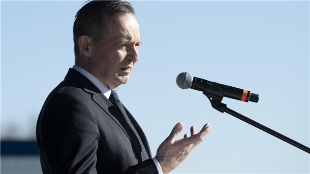 Ein Mann im Anzug steht vor einem Mikrofon und gestikuliert mit der rechten Hand. 