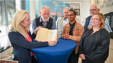 Menschen haben sich um einen Tisch mit blauer Decke versammelt, eine blonde Frau hat ein Buch aufgeschlagen und zeigt es der Kamera. 