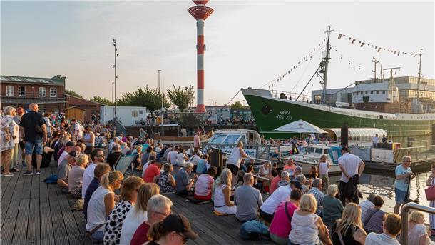 Blick auf den Hafen im Schaufenster Fischereihafen. Zu sehen sind das Museumsschiff "Gera" und das Oberfeuer sowie sitzende Menschen auf der Holzterrasse.