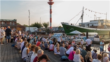 Blick auf den Hafen im Schaufenster Fischereihafen. Zu sehen sind das Museumsschiff "Gera" und das Oberfeuer sowie sitzende Menschen auf der Holzterrasse.