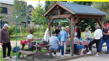 Eine Gruppe von Menschen sitzt und steht an zwei Holztischen mit Bänken. Nur einer der Tische ist überdacht.