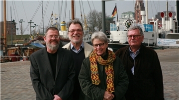 Drei Männer und eine Frau, alle zwischen Ende 50 und Anfang 70, blicken in die Kamera. Sioe stehen vor einem Hafenbecken, im Hintergrund sind alte Segelschiffe zu erkennen.