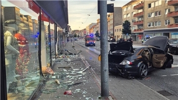 Glasscherben von einem Schaufenster liegen auf einem Fußweg. Daneben steht ein stark beschädigter schwarzer Wagen.