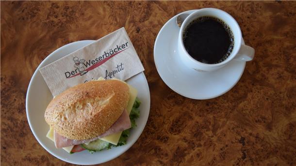 Es ist ein Brötchen zu sehen, das auf einem Teller angerichtet wurde. Daneben steht ein Becher Kaffee. 