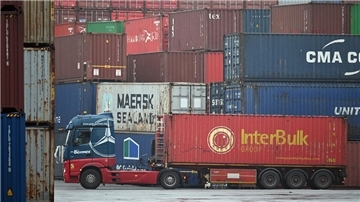 Ein Sattelzug mit einem roten Container der Marke 'InterBulk Group' steht auf einem Hafengelände, umgeben von gestapelten Frachtcontainern in verschiedenen Farben und mit Markenaufdrucken wie 'Maersk Sealand' und 'CMA CGM'.