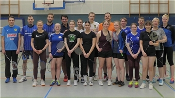 Eine Gruppe mit Sportklamotten bekleidet und Badmintonschläger in den Händen steht in einer Turnhalle. Alle lächeln in die Kamera. 