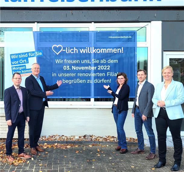 Fünf Bankmitarbeiter weisen auf ein großes Plakat mit der Aufschrift "Herzlich willkommen! Wir freuen uns, Sie ab dem 3. November 2022 in unserer renovierten Filiale begrüßen zu dürfen."