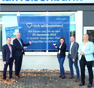 Fünf Bankmitarbeiter weisen auf ein großes Plakat mit der Aufschrift "Herzlich willkommen! Wir freuen uns, Sie ab dem 3. November 2022 in unserer renovierten Filiale begrüßen zu dürfen."