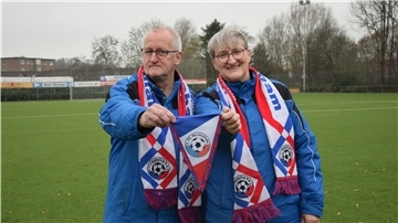 Ein Mann und eine Frau stehen auf einem Fußballfeld. Beide tragen Fan-Schals eines Vereins. Der Mann zeigt einen Wimpel des Vereins in die Kamera.