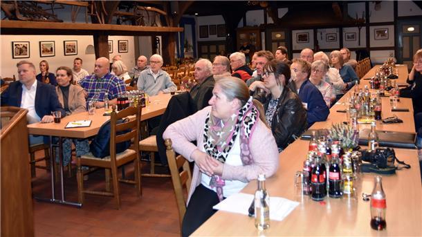 Etwa 30 Menschen sitzen an langen Tischreihen und blicken alle in eine Richtung.