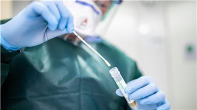 Canan Emcan, Stationsleiterin auf der Infektionsstation der Uniklinik Essen, steht in Schutzkleidung und mit einer Atemmaske in einer Schleuse und hält ein Abstrichröhrchen in den Händen.