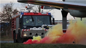 Ein rotes Flughafenlöschfahrzeug mit eingeschaltetem Blaulicht fährt durch dichte Rauchwolken in Gelb und Rot. Im Vordergrund ist der Rauch stark zu sehen, im Hintergrund Bäume und ein Teil eines Flugzeuges.