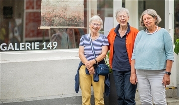 Drei ältere Frauen stehen vor einer Galerie und blicken in die Kamera.