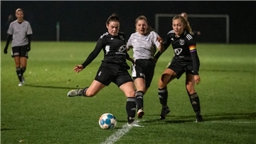 Drei Frauen in Sporttrikots laufen um einen Fußball herum.