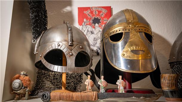 Zwei eiserne Helme, die wirken, als kämen sie aus dem Mittelalter. Unter ihnen sind vier fingergroße Figuren platziert, die ebenso an das Mittelalter erinnern.