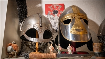 Zwei eiserne Helme, die wirken, als kämen sie aus dem Mittelalter. Unter ihnen sind vier fingergroße Figuren platziert, die ebenso an das Mittelalter erinnern.