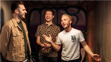 Drei Mitglieder der Band „Von Grambusch“ lachen und unterhalten sich gut gelaunt vor einer farbig besprühten Wand.