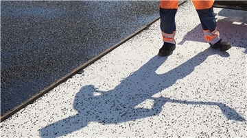 Der Schatten eines Bauarbeiters fällt auf den Boden einer Straße, die asphaltiert wird. Die Kreisstraße 48 im Landkreis Cuxhaven wird in zwei Abschnitten saniert und bleibt bis September gesperrt.