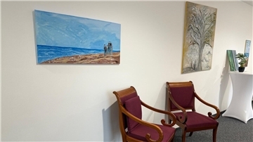 Kunstwerke hängen an einer Wand, davor stehen zwei Stühle. Die Künstlerin Käthe Goltz stellt 27 farbenfrohe Werke im Amtsgericht Zeven aus.