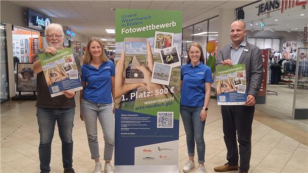 Zwei Frauen und zwei Männer stehen im Gang eines Einkaufszentrums mit Aufsteller und Plakaten zu einem Fotowettbewerb 