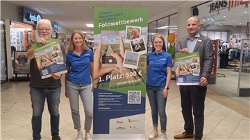 Zwei Frauen und zwei Männer stehen im Gang eines Einkaufszentrums mit Aufsteller und Plakaten zu einem Fotowettbewerb 