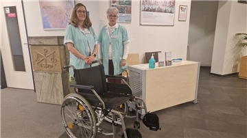 Zwei Frauen stehen mit einem Rollstuhl in der Eingangshalle eines Krankenhauses. 