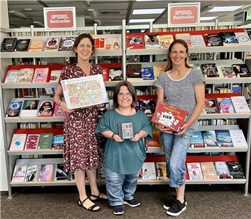 Drei Frauen stehen vor einem Regal und halten Bücher und Spiele in der Hand.