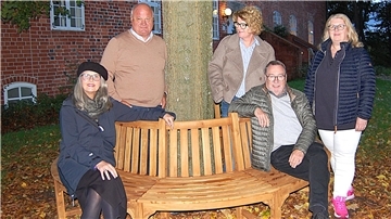 Fünf Personen stehen und sitzen um eine halbrunde Holz-Baumbank im herbstlich gefärbten Burghof. Im Hintergrund ist ein historisches Backsteingebäude zu sehen.